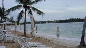 A beach with white lounge chairs and umbrellas, palm trees, and a view of the ocean. - Punta Aguila Spectacular 4 Bdr / 6 Bath Free Golf Cart  Chef and Waiter Included (La Romana)