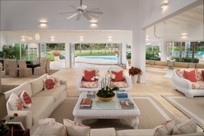 A spacious living area with a large window overlooking a swimming pool, furnished with white sofas and a central coffee table.