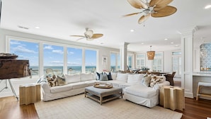 A spacious living room with a large window overlooking the ocean, a ceiling fan, and a dining area with a view. - 6 BR + Bonus Room, Luxury Oceanfront, Pets ok, Elevator, 2 Living Rooms - The Loggerhead (Folly Beach)