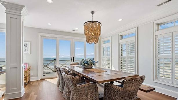 A dining area with a wooden table, wicker chairs, a chandelier, and large windows with ocean views. - 6 BR + Bonus Room, Luxury Oceanfront, Pets ok, Elevator, 2 Living Rooms - The Loggerhead (Folly Beach)