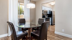 A dining area with a glass table and four chairs, a kitchen in the background, and a view of the outdoors.