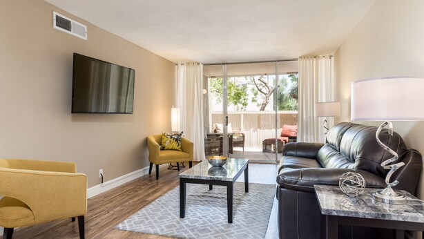 A living room with a flat-screen TV, a yellow armchair, a black leather sofa, a marble coffee table, and a view through a sliding glass door.