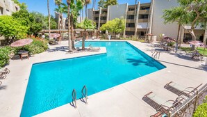 A swimming pool with lounge chairs and umbrellas, surrounded by palm trees and a multi-story building.