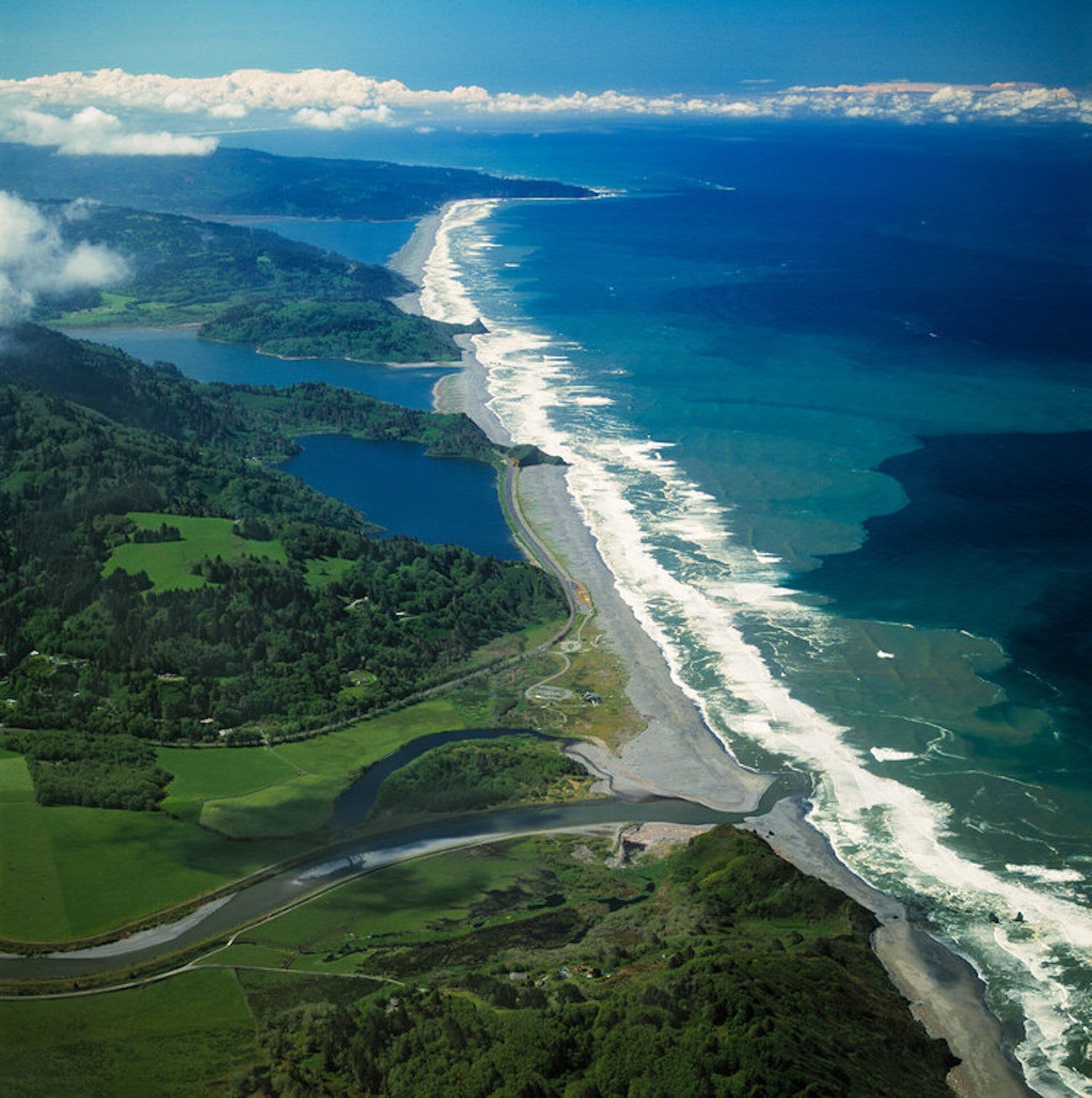 Vista aérea de una zona costera con un camino serpenteante, campos verdes y una playa.