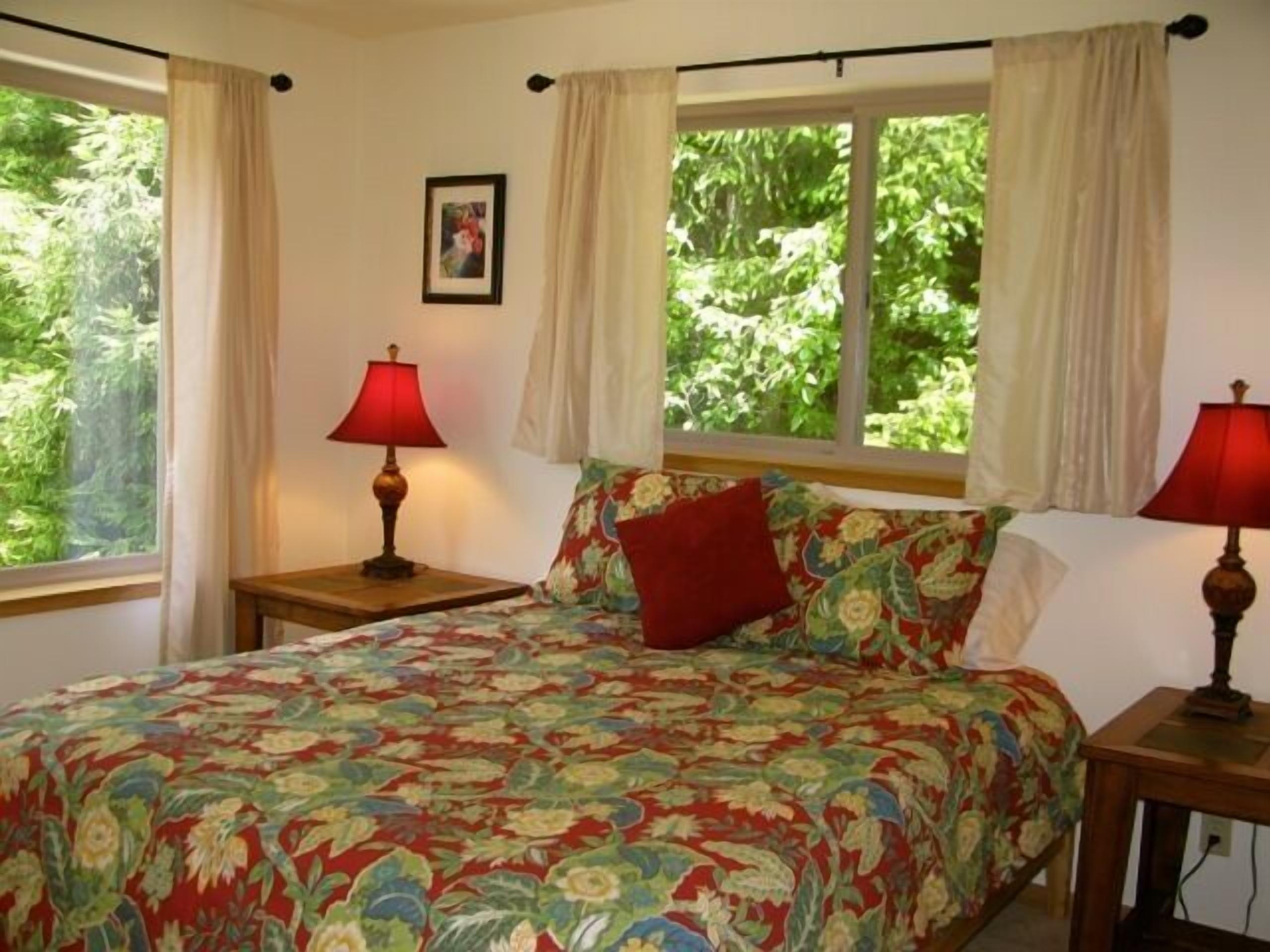 Une chambre à coucher avec une couvre-lit à motifs floraux, deux lampes de chevet, une fenêtre avec des rideaux et un tableau encadré accroché au mur.