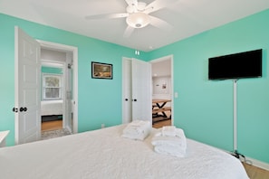 Un dormitorio con cama, toallas blancas, ventilador de techo, televisor y puerta que da a otra habitación.