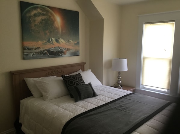 A neatly made bed with a white duvet and a dark throw blanket, a wooden headboard, a bedside lamp, and a painting of a celestial scene on the wall.