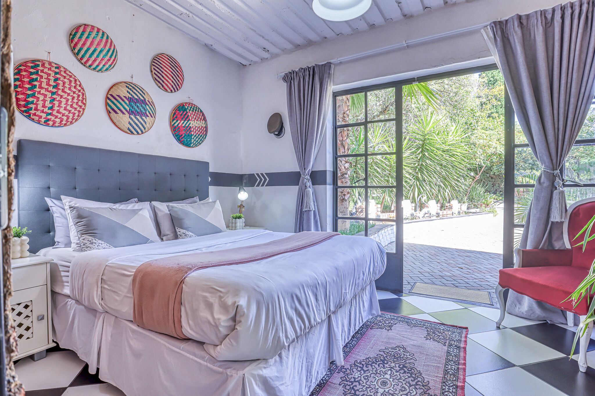 A bedroom with a large bed, a nightstand, a red chair, and a view of the outdoors through a window.