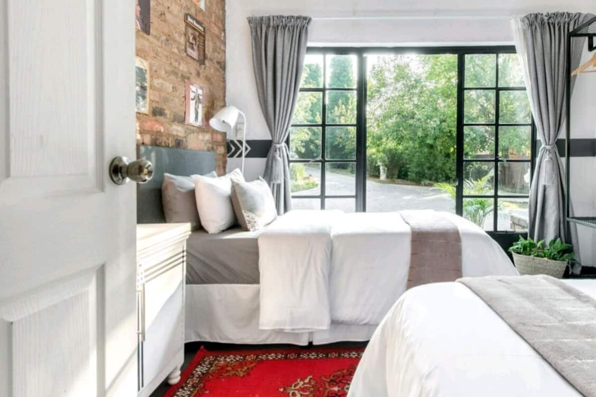 A bedroom with a brick wall, a large window with curtains, a bed with white bedding, and a red patterned rug.