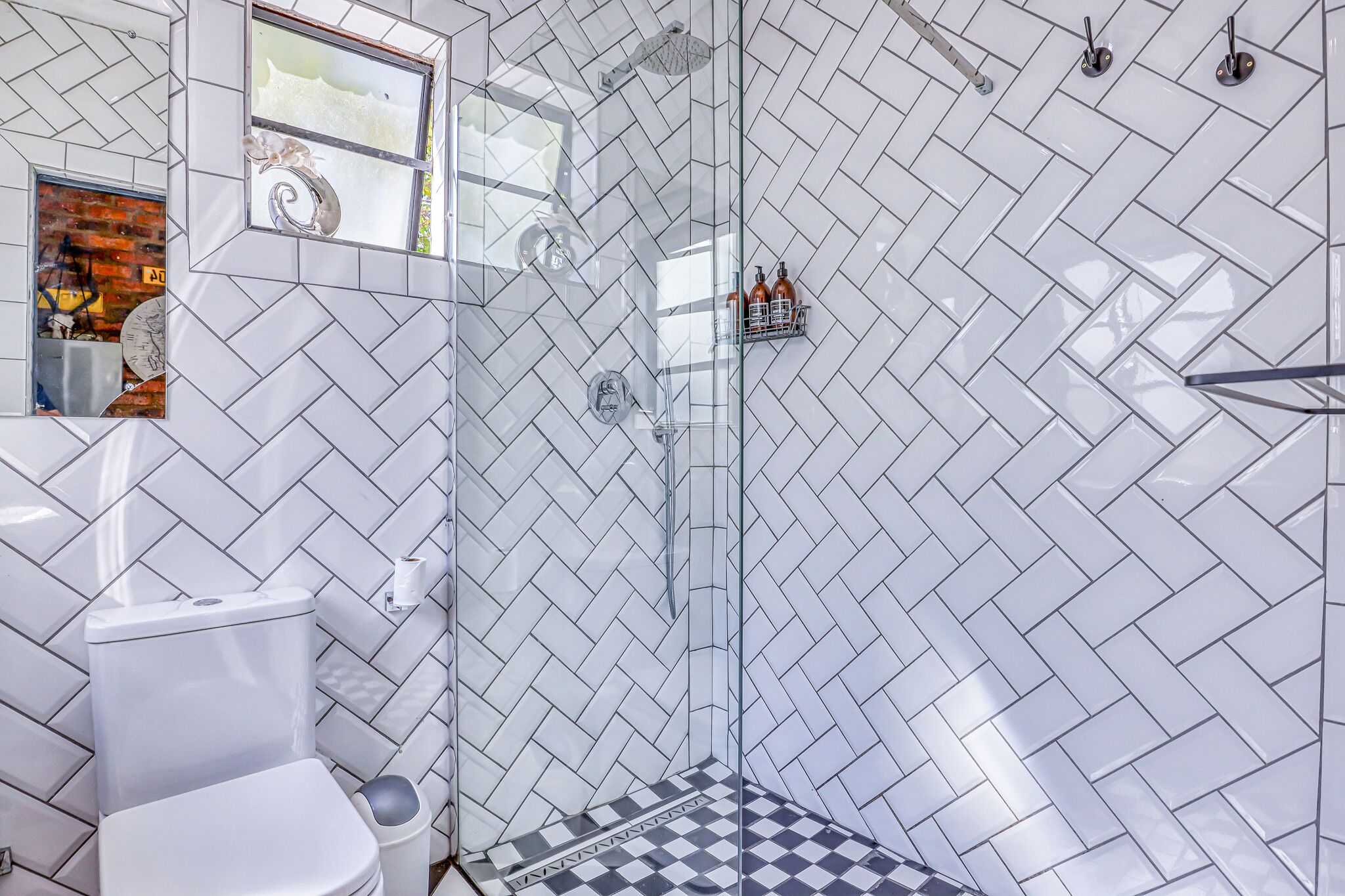 Un baño con azulejos blancos en patrón de espiga, un box de ducha de vidrio y piso a cuadros.