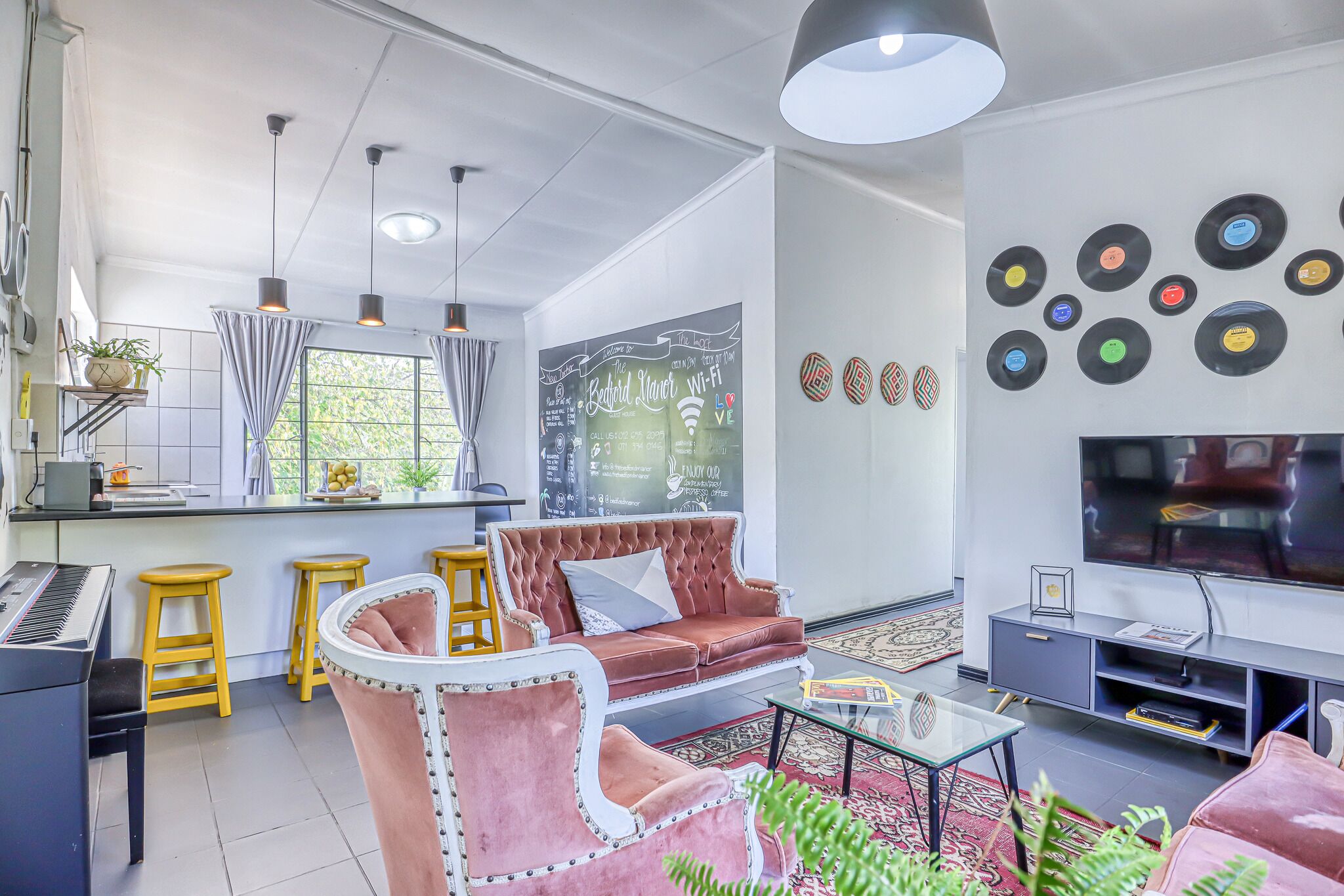 A modern living room with a sofa, coffee table, and a TV. There is a kitchen area with a piano, stools, and a chalkboard wall.