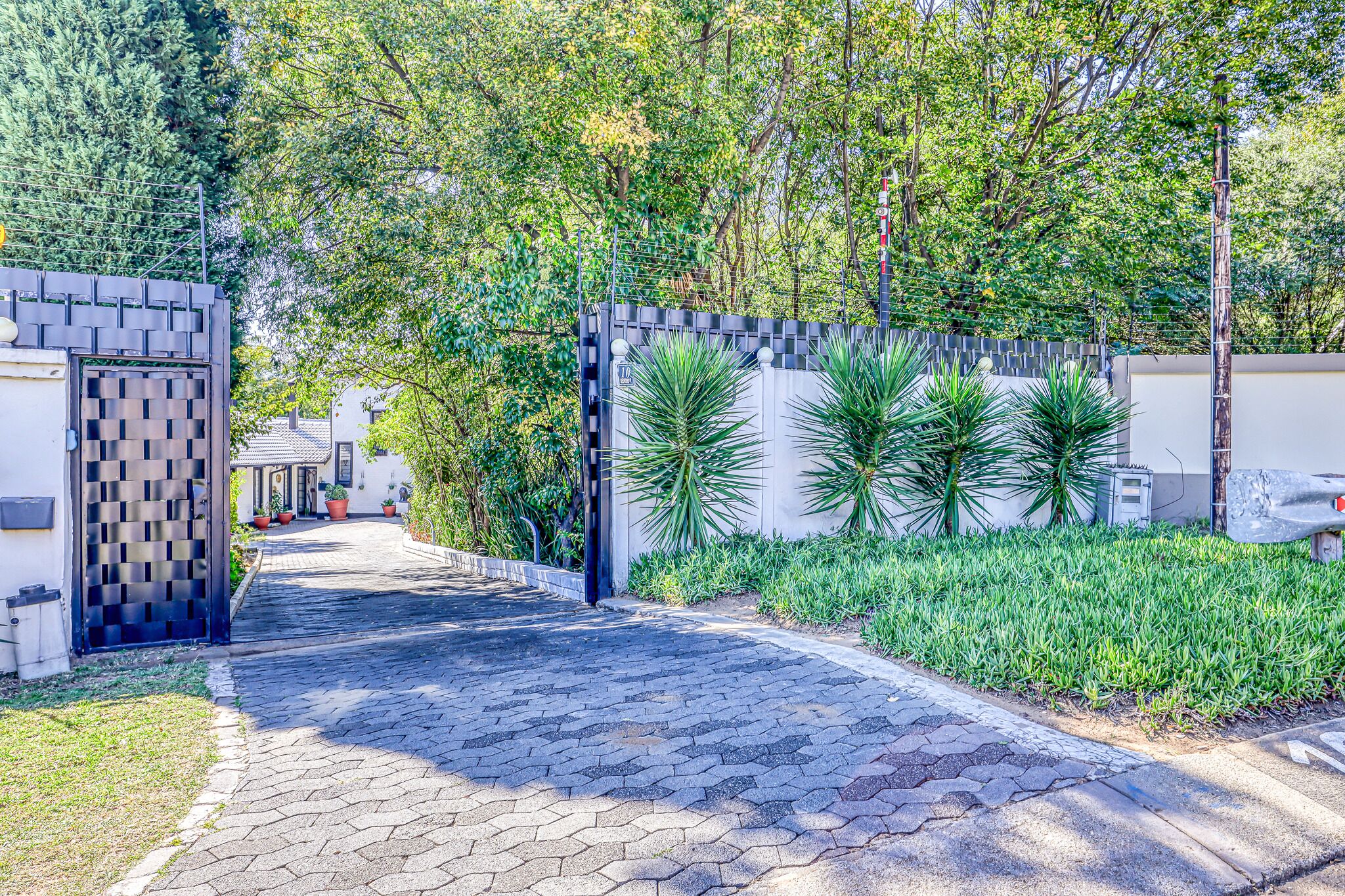 Un sendero pavimentado que conduce a una entrada porticada de diseño moderno, rodeada de abundante vegetación y un edificio blanco.