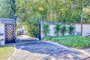 Un sendero pavimentado que conduce a una entrada porticada de diseño moderno, rodeada de abundante vegetación y un edificio blanco.