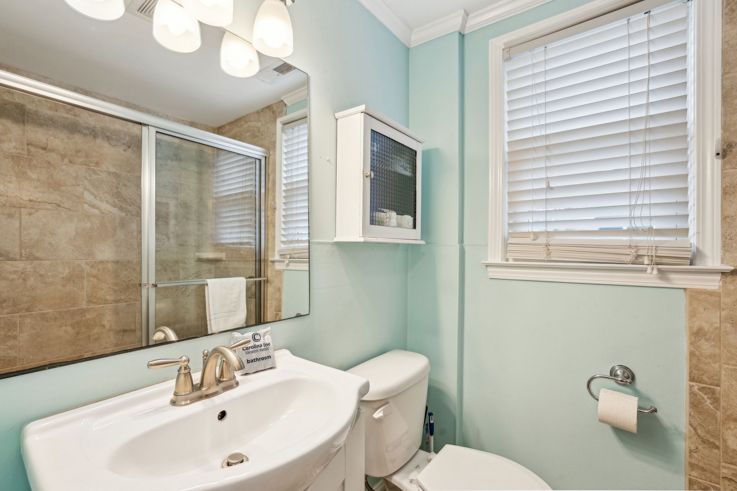 A bathroom with a white sink, toilet, and a mirror. There is a window with blinds and a towel rack.