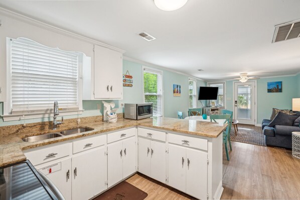 A kitchen with white cabinets, a stainless steel sink, and a microwave. There is a dining area with a table and chairs. - Renovated & Only 2.5 Blocks to Shops & Restaurants (Folly Beach)
