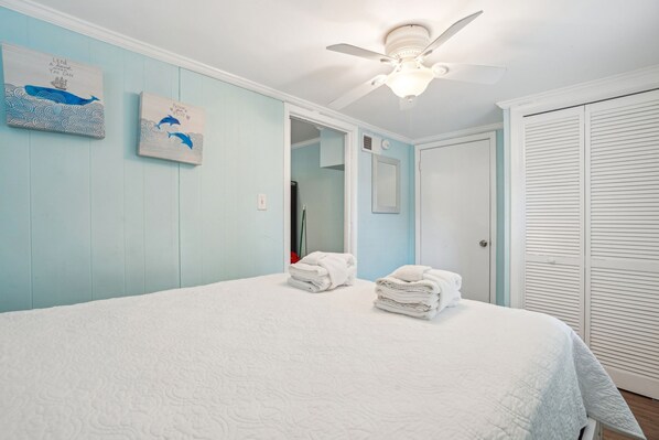 A bedroom with a bed, two wall-mounted artworks, a ceiling fan, a closet, and a door leading to another room. - Renovated & Only 2.5 Blocks to Shops & Restaurants (Folly Beach)