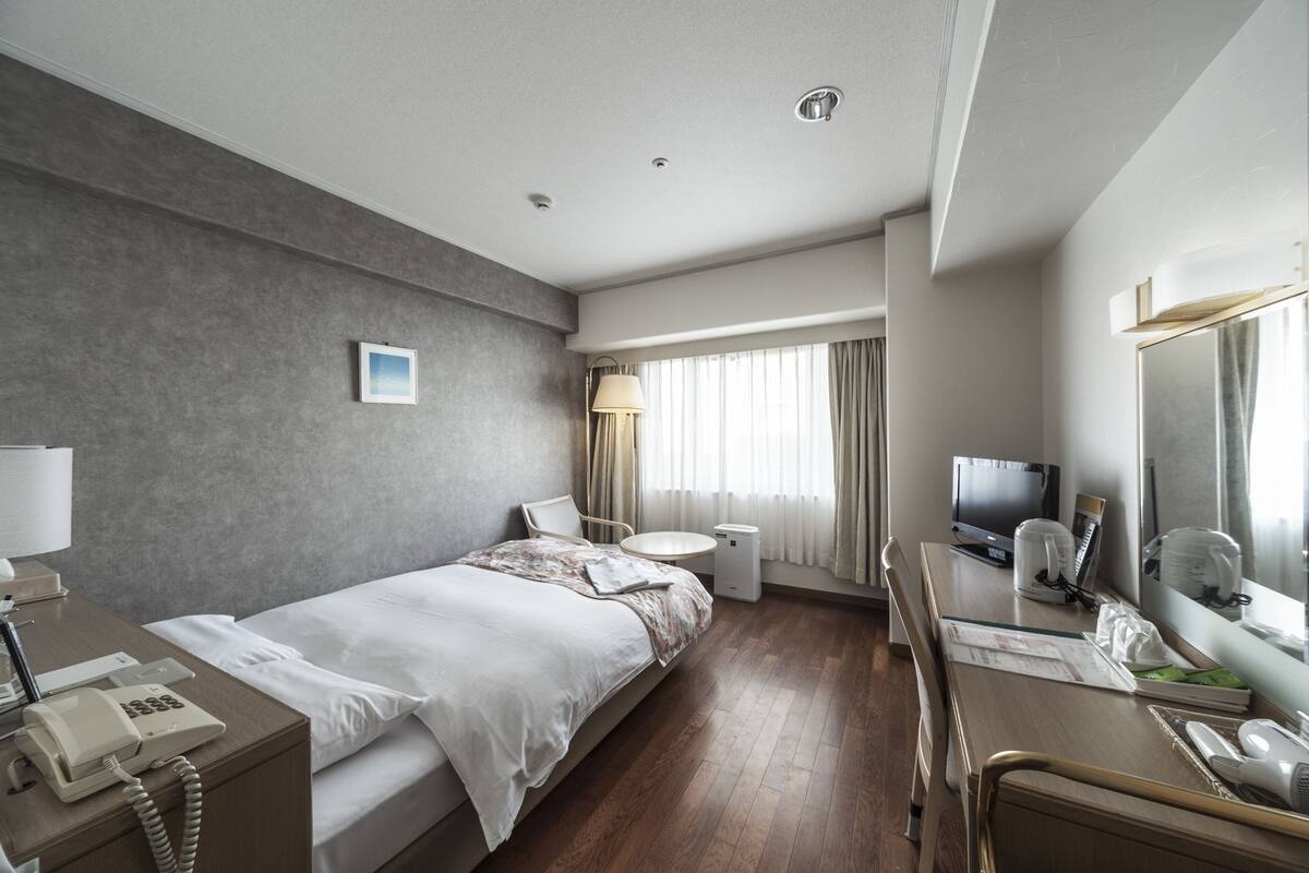 A modern hotel room with a bed, bedside table, desk, chair, and a large mirror.