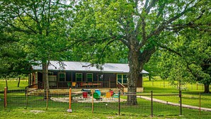 A rustic building with a porch, surrounded by lush greenery and a fenced playground area. - Panoramic Country Views ~ Wildlife ~ Butterflies & Birds ~ Star-gazing ~ Hiking (Leakey)