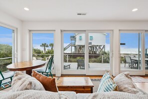 A spacious living area with a wooden table and chairs, a sofa with cushions, and large sliding glass doors leading to an outdoor deck with a view of a beach and a white building. - NorthStar! FREE $250 Beach Gear Credit! Ocean Views! Open floorplan w/Mother-In-Law Suite (Folly Beach)
