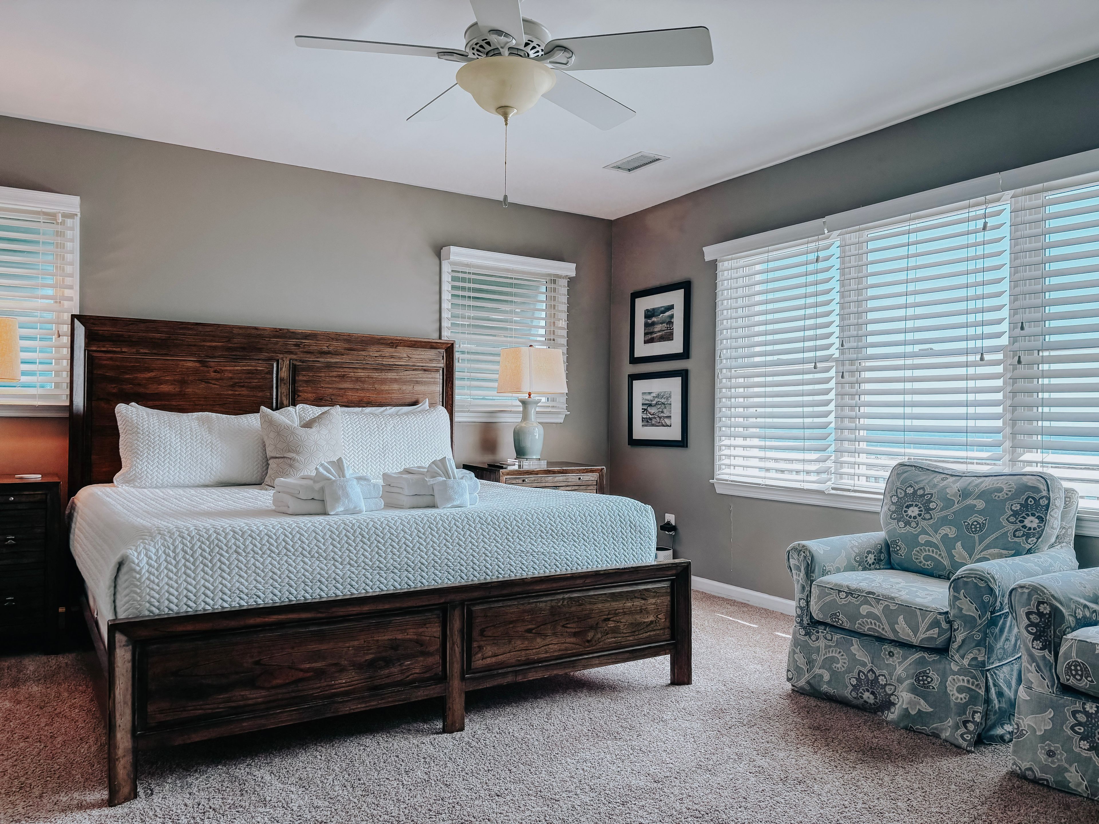 Une chambre à coucher avec un lit en bois, un ventilateur de plafond et une vue sur l’extérieur grâce à de grandes fenêtres.