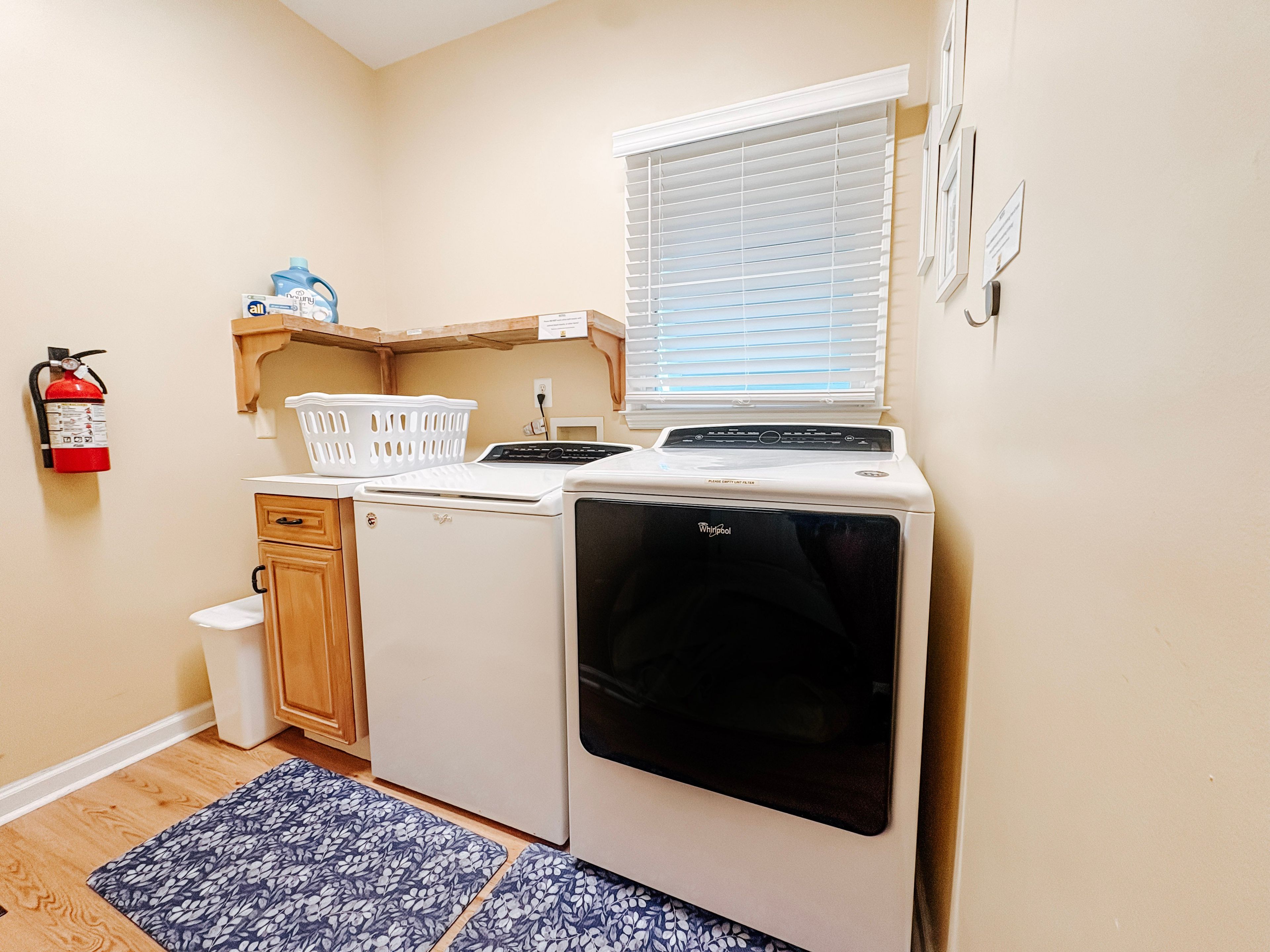 A compact kitchen with a washing machine, a dryer, a small window with blinds, a fire extinguisher, and a laundry basket on a shelf.