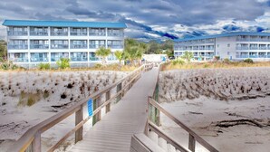 Ein hölzerner Steg führt zu einem Strandhotel mit mehreren Balkonen und einem blauen Dach.