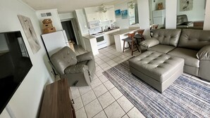 Ein modernes Wohnzimmer mit Sofa, Hocker und Fernseher. Es gibt eine Küchenzeile mit Kühlschrank, Mikrowelle und Schränken.