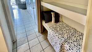 Ein schmaler Flur mit einem Etagenbett auf der rechten Seite, einer Tür mit einem Kissen mit Strandmotiv und einem gerahmten Bild an der Wand.