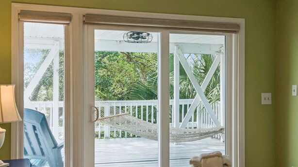 A room with a bed, a chair, and a view of a wooden deck and palm trees through a sliding glass door. - Ocean, Marsh, & River Views! Dock For Fishing & Sunsets (Folly Beach)