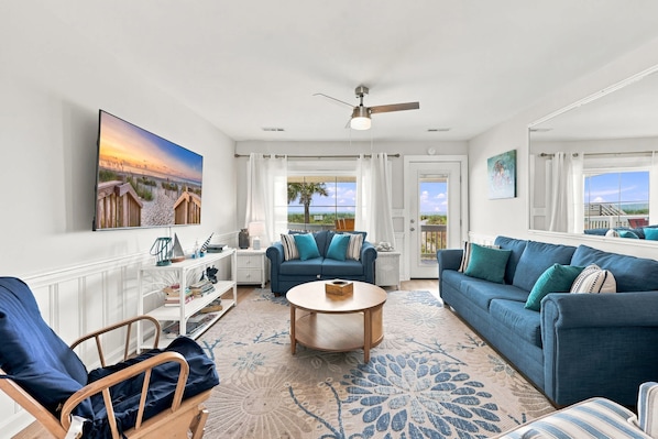 A spacious living room with a large painting, a ceiling fan, and a view of the beach through the windows.