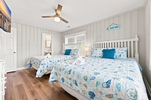 Un dormitorio con dos camas, ventilador de techo, ventana con persianas y un letrero que indica que es una casa de playa.