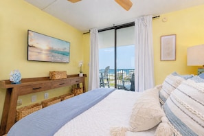 A bedroom with a bed, a TV, a balcony with a view, and a wall with a framed picture.