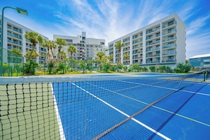 A tennis court with a net in the foreground, surrounded by a fence, and a multi-story hotel building in the background.