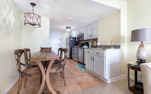 Una cocina con armarios blancos, microondas y refrigerador. Hay una mesa de comedor con sillas y una araña.