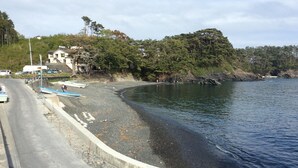 A coastal road with boats docked on the shore, a building with a sloped roof, and dense forest in the background. - Umisanpo (Ofunato)