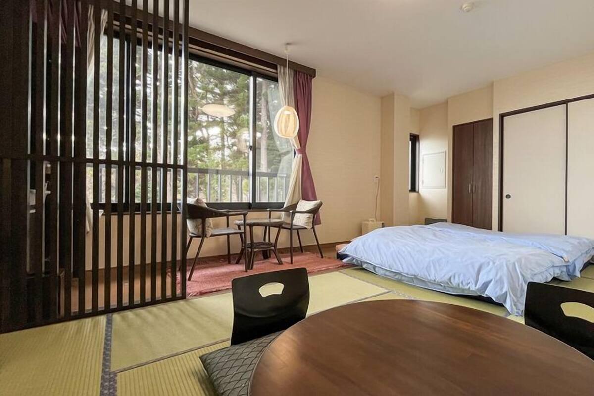 A room with a bed, a desk, and a chair, with a sliding door and a view of trees outside.