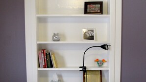 Un lit bien fait, avec une literie à rayures, une bibliothèque blanche remplie de livres et d’objets décoratifs, et une petite lampe de table.