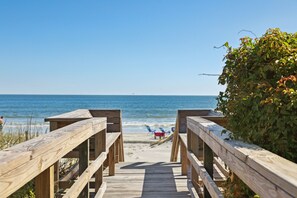 A wooden boardwalk leading to a beach with a clear view of the ocean and a few beach chairs.