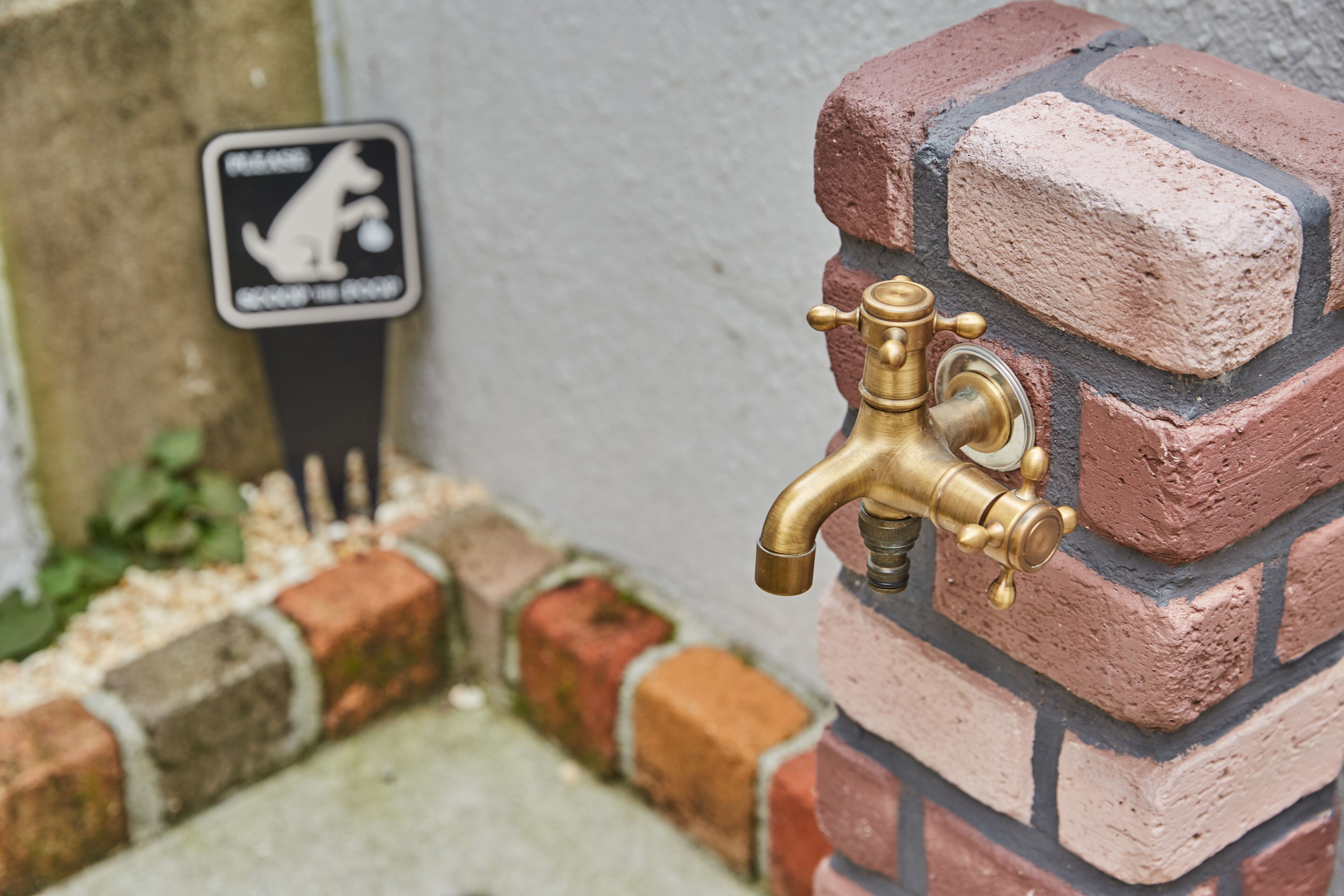 Ein Messing-Außenhahn an einer Ziegelwand, mit einem Schild, das auf ein hundefreundliches Gebiet in der Nähe hinweist.