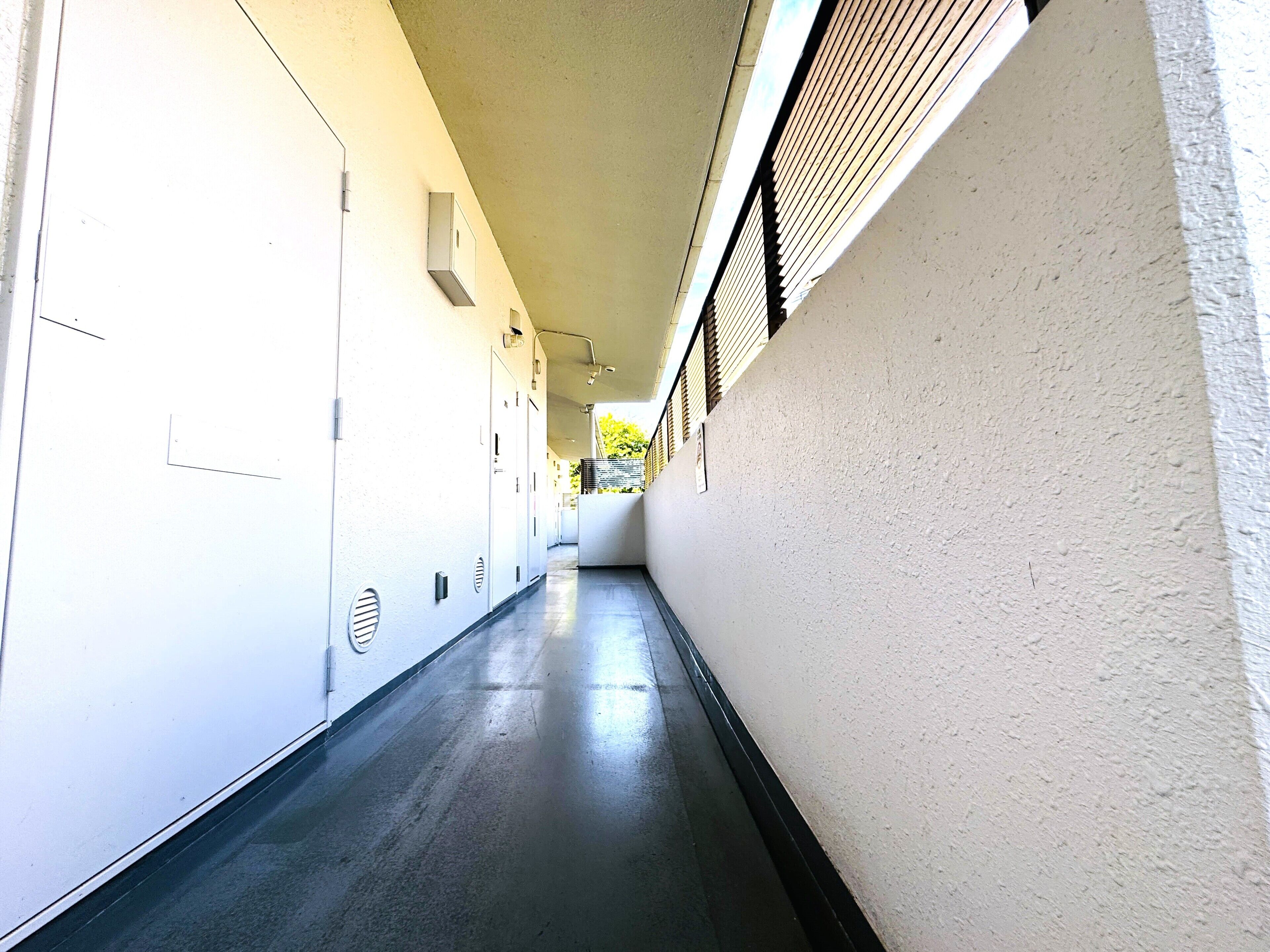 A long, narrow hallway with white walls and a dark floor. There are doors on the left side and a window with blinds on the right.