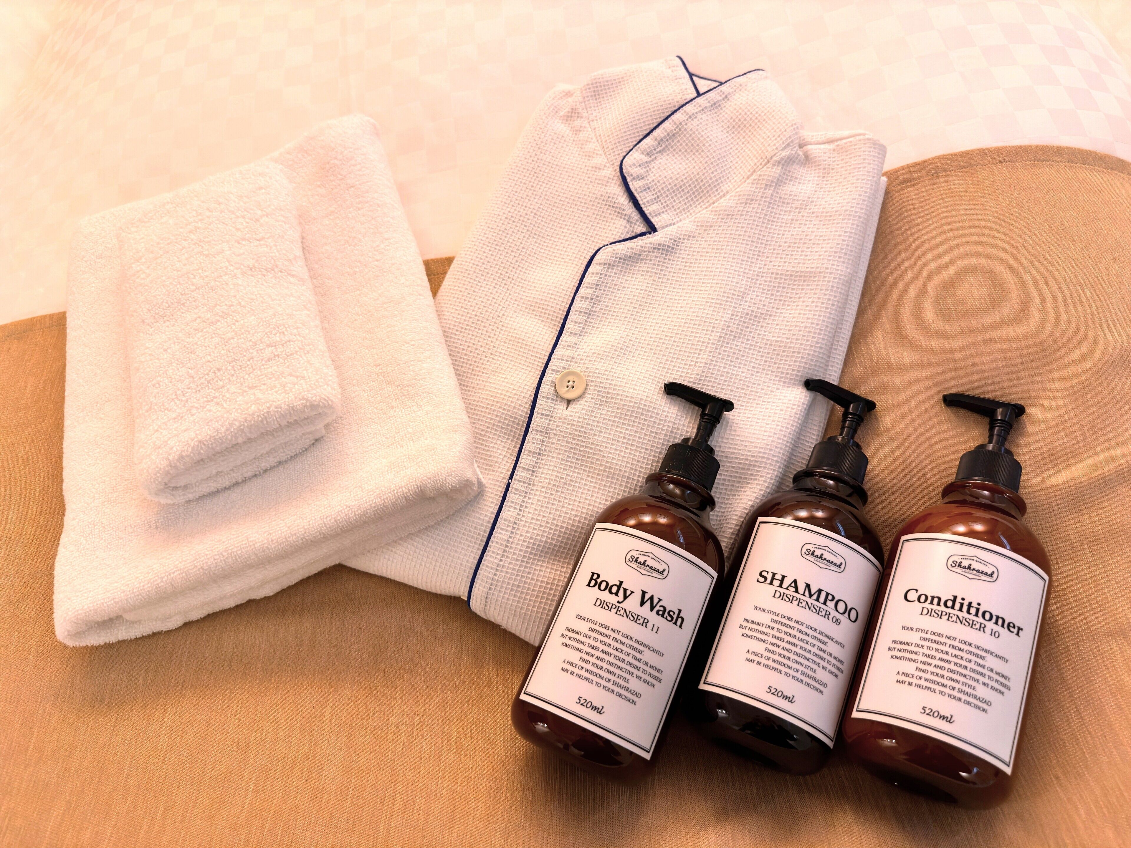 Une serviette blanche soigneusement pliée, un peignoir à rayures et trois flacons de produits de soins pour la peau (gel douche, shampoing et après-shampoing) sont posés sur une surface en bois.
