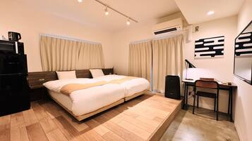 A modern hotel room with a large bed, a desk with a chair, a black speaker, and a wall-mounted air conditioner.