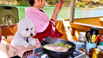 A person sitting at a table with a dog, a portable stove with food, and a scenic view of a body of water.