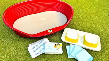 A red pet bed, two yellow and one blue water dish, and a pack of baby wipes on a grassy surface.