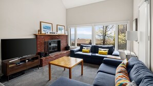 A living room with a TV, fireplace, sofa, and large windows. - 41 Summit Condo Near Mountain Shuttle by RedAwning (Warren)