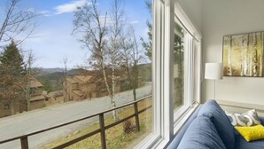 A room with a large window overlooking a street and buildings, a blue sofa with a yellow pillow, and a painting on the wall. - 41 Summit Condo Near Mountain Shuttle by RedAwning (Warren)