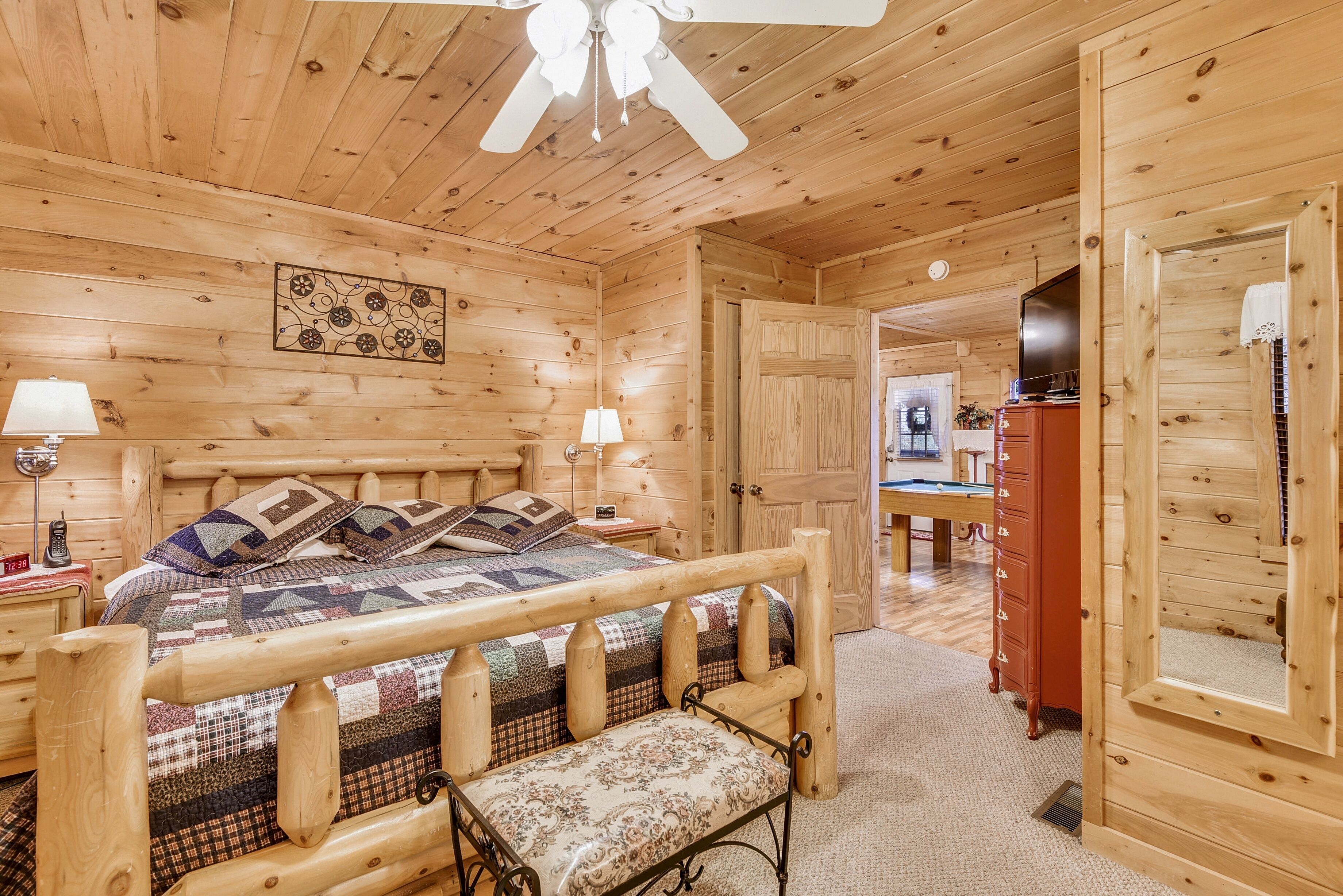 A wooden cabin bedroom with a bed, bedside lamps, a bench, a ceiling fan, and a door leading to another room.