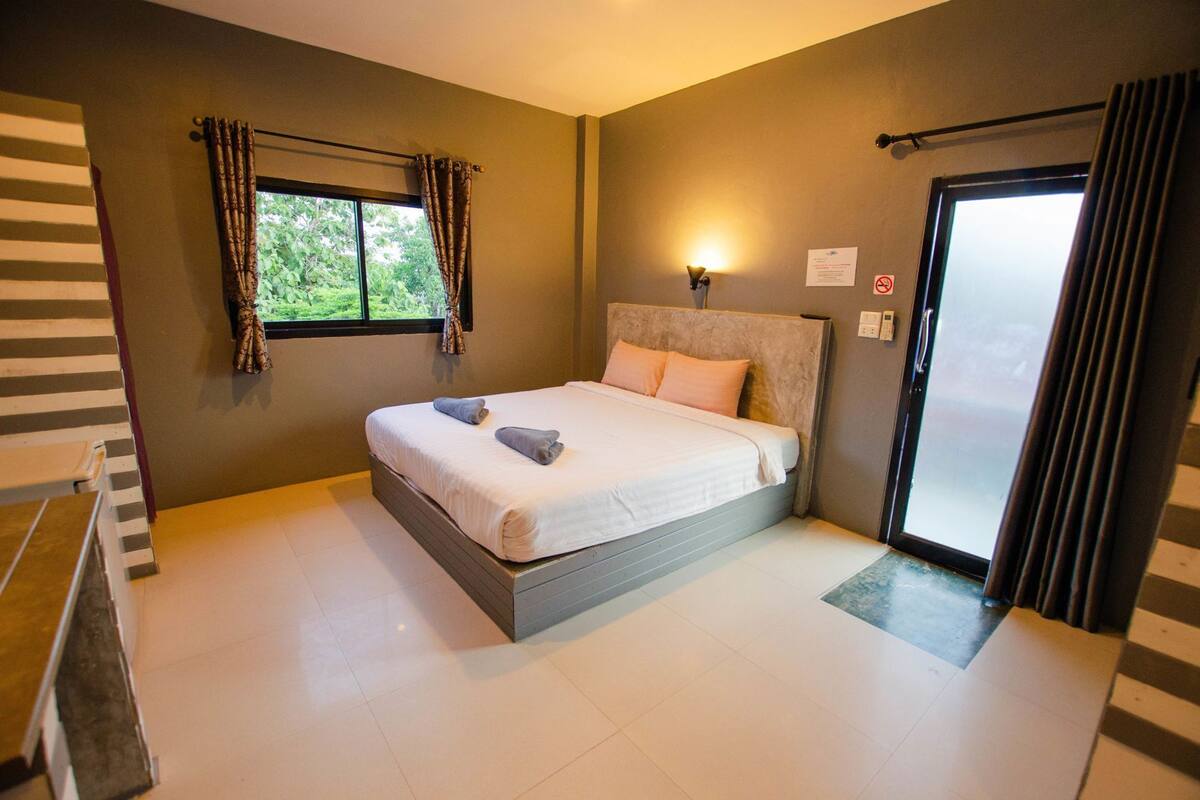 Um quarto com uma cama, uma janela com cortinas, uma mesinha de cabeceira e uma porta.