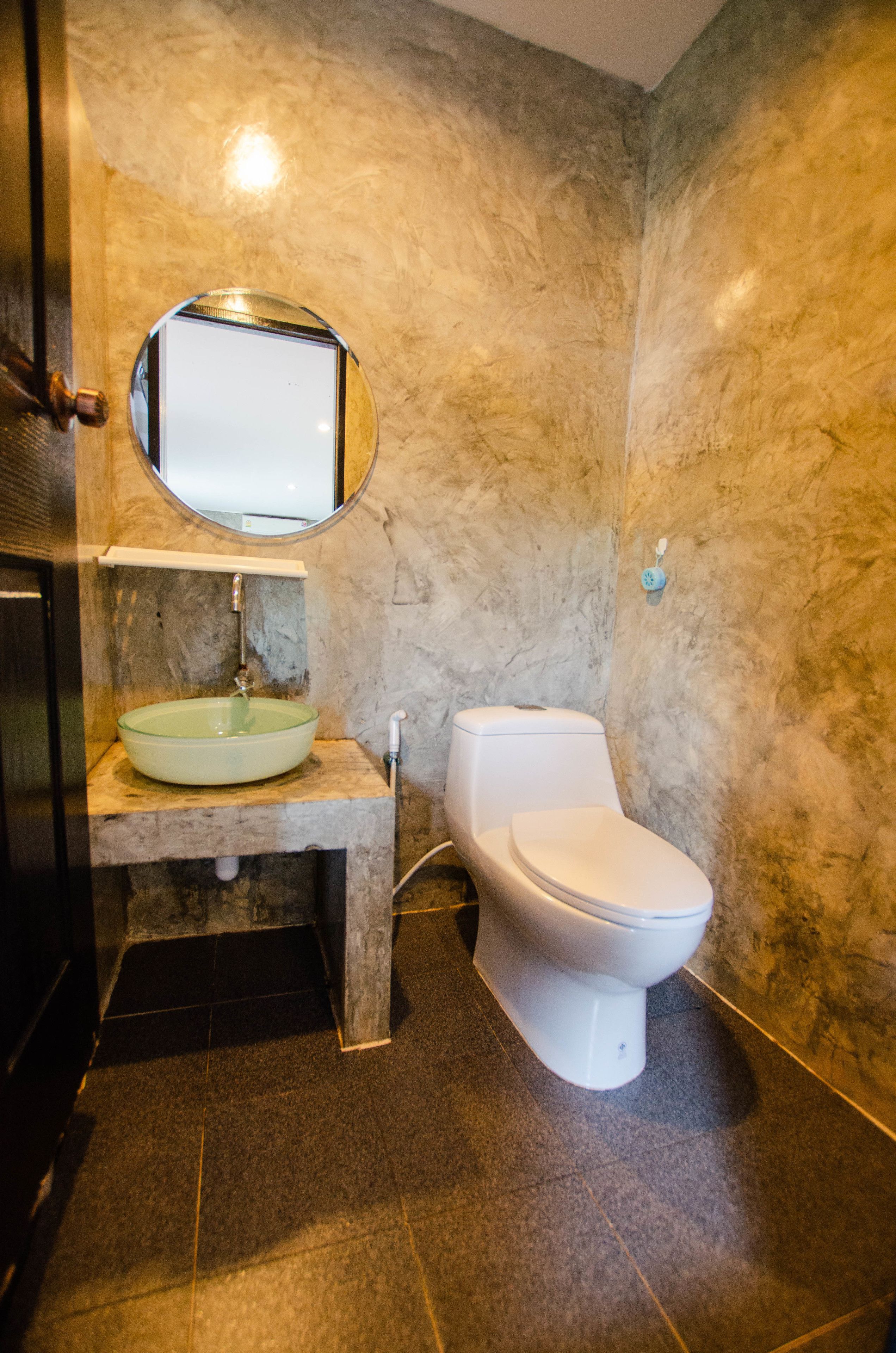 Un baño con pared de mármol, un espejo redondo, un lavabo de vidrio y un inodoro blanco.