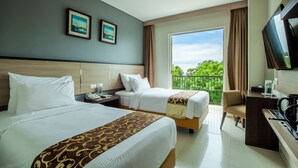Habitación de hotel con dos camas, un escritorio, televisor y balcón con vistas a la vegetación.